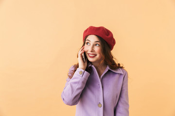 Portrait of young girl 20s in coat and hat using smartphone for call, while standing isolated over beige background