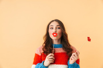 Image of european woman 20s with long hair smiling and holding paper lips, standing isolated over beige background