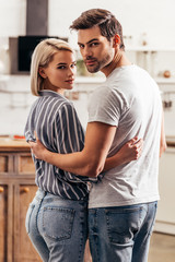 selective focus of handsome boyfriend and attractive girlfriend hugging and standing in kitchen while looking at camera