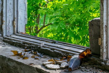 view of the broken window of the destroyed old building with broken bottles on the windowsill, behind which you can see the green thickets of trees