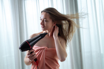 Beautiful young brown haired woman in bath towel is using a hair dryer for drying her long hair after shower in room. Hair Care.
