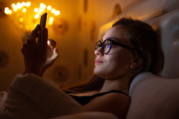 Young, glasses woman before bedtime chatting with loved people and friends. Chat night and surfing phone. Social networks and phone addiction.