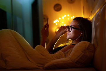 Young, glasses woman in headphones before bedtime listening to relaxing music and chatting with loved people and friends. Chat night and surfing phone. Social networks and phone addiction.