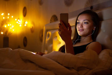 Young woman in headphones before bedtime listening online to relaxing music and chatting. Chat night and surfing phone. Social networks and phone addiction.