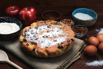Apple pie made from flour, apples, raisins, lemon zest, honey, sugar and walnuts on a brown wooden board. Next to eggs, cinnamon, raisins, apples and honey.