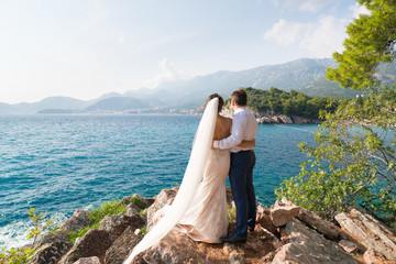 the newlyweds stand on the rocks and look at the sea holding hands