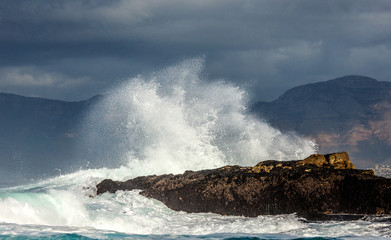 Naklejka na ściany i meble Big waves break on the rocks in the sea against the backdrop of the coastline. Beautiful seascape. A beautiful moment. Very dynamic photo. Cape Town. False Bay. South Africa.