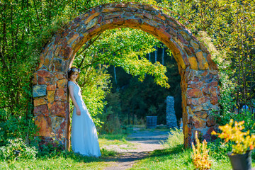 beautiful and young bride in white dress and circlet of flowers posing in stone arch