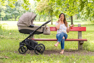 A smiling young mother sitting on a bench next to a stroller in the park and talking on her phone.