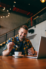Smiling bearded freelancer mixing espresso with spoon and holding in other hand smart phone. Laptop on the table, cafeteria interior.