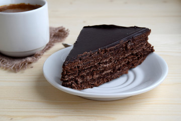 The piece of chocolate cake  is on a white plate. The cup with coffee is near cake.