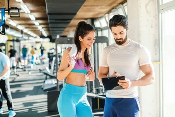 Tuinposter Fit smiling Caucasian woman with towel around neck looking at clipboard. Her personal trainer writing and showing her result of training. Gym interior. © dusanpetkovic1