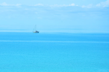 Fototapeta na wymiar A distant, solitary, traditional, cutter rigged sailboat sitting at anchor in the Bahamas on a calm Caribbean blue sea that stretches out into a blue cloud sky with no visible horizon. Room for copy.