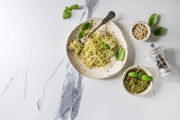 Classic italian spaghetti pasta with pesto sauce, pine nuts, olive oil and fresh basil. Served in ceramic plate with fork and ingredients above over white marble background. Flat lay, space