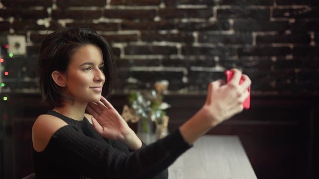 Young girl is sitting at the cafe.She has short hair and always fix it with hand. Also she is making selfie and smiling on camera.