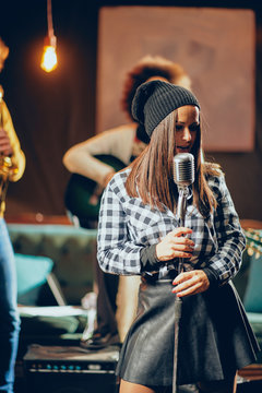 Portrait of young talented rocker girl singing and holding microphone in home studio. In background bands playing instruments.
