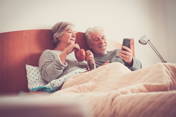Cheerful caucasian senior adult couple at home enjoying breakfast together at bed during a new morning life - real people living forever married in love enjoying technology internet phone