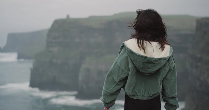 A cute small brunette lady in front of the camera on the top of Cliffs of Moher she impressed of place moving slow in front of the camera , and turn with face to the ocean view.