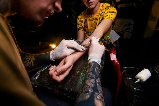 Handsome young guy in a black hat and with tattoos, beats a tattoo on his arm, tattoo salon, tattoo artist