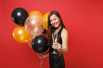 Attractive young girl in little black dress celebrating, holding glass of champagne, air balloons isolated on red background. St. Valentine's Day, Happy New Year birthday mockup holiday party concept.