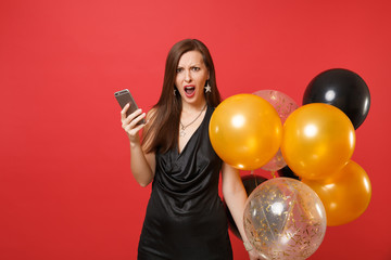 Irritated young girl in black dress swearing holding air balloons using mobile phone while celebrating isolated on red background. Valentine's Day Happy New Year birthday mockup holiday party concept.