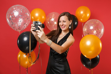 Joyful young girl in little black dress doing taking selfie shot on retro vintage photo camera on bright red background air balloons. Women's Day, Happy New Year birthday mockup holiday party concept.