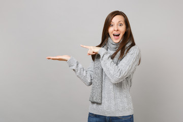 Amazed young woman in sweater, scarf pointing hand index finger aside keeping mouth open isolated on grey background. Healthy fashion lifestyle people emotions cold season concept. Mock up copy space.