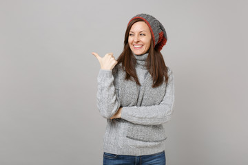 Cheerful young woman in gray sweater, hat scarf looking, pointing thumb aside isolated on grey background. Healthy fashion lifestyle, people sincere emotions, cold season concept. Mock up copy space.