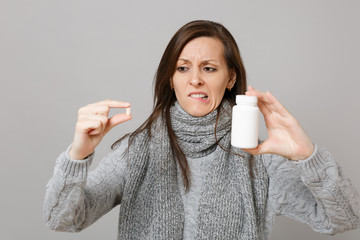 Puzzled young woman in gray sweater, scarf biting lips holding medication tablets aspirin pills in bottle isolated on grey background. Healthy lifestyle ill sick disease treatment cold season concept.