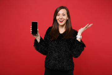 Naklejka premium Amazed young woman in black fur sweater spreading hand, holding mobile phone with blank empty screen isolated on bright red background. People sincere emotions, lifestyle concept. Mock up copy space.