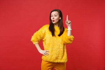 Pretty young woman in yellow fur sweater holding index finger up with great new idea isolated on bright red wall background in studio. People sincere emotions, lifestyle concept. Mock up copy space.