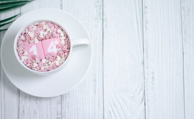 Fototapeta na wymiar Pink square wood with white numbers 1 and 4.Gray square wood with white letters FEB.February 14 Valentine's Day.White coffee cup on wooden floor.Many small hearts, white and pink