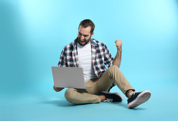Emotional young man with laptop celebrating victory on color background