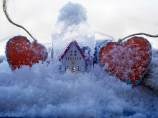 A small white toy house with a red roof and a lighted illumination and a pair of orange felt hearts on the snow covered with snow, the concept of winter seasonal holidays, Valentine's Day greetings, h