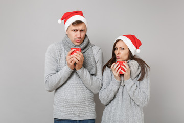 Fun couple girl guy in red Santa Christmas hat gray sweaters scarves hold cups of tea isolated on grey wall background, studio portrait. Happy New Year 2019 holiday party concept. Mock up copy space.