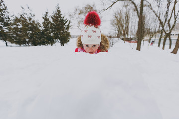 Funny little girl in winter warm clothes and hat hiding, peeping out from snowball in snowy park or forest outdoors. Winter fun, leisure on holidays. Love relationship family people lifestyle concept.