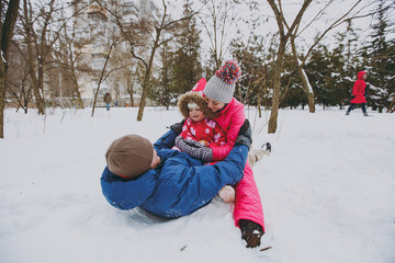 Funny family woman, man and little girl in warm clothes lying, sitting on snow in park or forest outdoors. Winter fun, leisure on holidays. Love childhood relationship family people lifestyle concept.