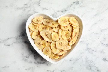 Heart shaped plate with banana slices on marble table, top view. Dried fruit as healthy snack