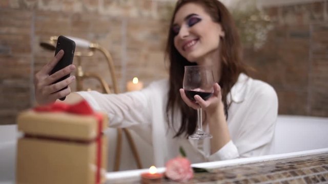 Young woman with bright makeup making selfie sitting in the bath with red wine glass. Cute girl relaxing in the bathroom. Focus moves from the background to the foreground and back.
