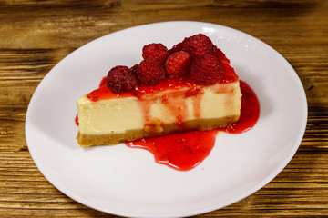 Piece of tasty New York cheesecake with raspberries and raspberry jam in a white plate on wooden table