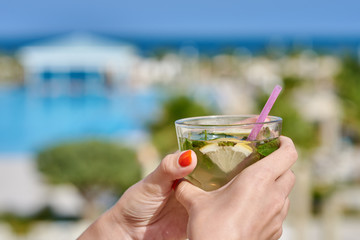 Woman is holding mojito cocktail in her hands against wonderful hotel’s view. Close-up photo.