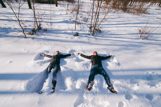 Joyful couple woman and man in winter warm clothes making snow angels while lying in city park or forest outdoors. Winter fun, leisure on holidays. Love relationship family people lifestyle concept.
