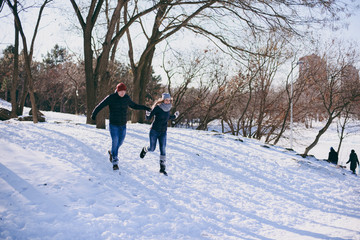 Cheerful young couple woman, man in warm clothes running, holding hands, walking in snowy city park or forest outdoors. Winter fun, leisure on holidays. Love relationship people lifestyle concept.