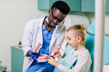 Little patient at the dentist's office. Young African American dentist talking with patient small boy holding artificial jaws in his hands.