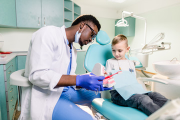 Fototapeta na wymiar Happy smiling black dentist tells Caucasian little boy how to brush his teeth. Caries prevention, Dentistry, teeth hygiene concept