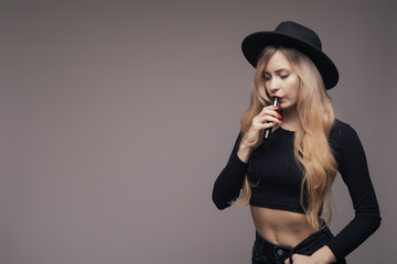 Attractive trendy woman in hat and black shirt vaping and blowing smoke on white background