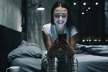 Dating app concept. Young pretty female sitting on bed at night, looking at smartphone, laughing, texting
