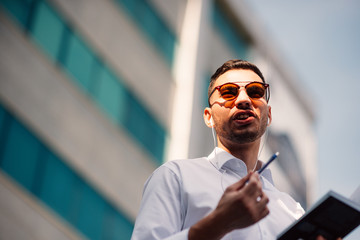 Confident businessman is in urban neighborhood with his headphones on, holding a pen and holding his notebook.