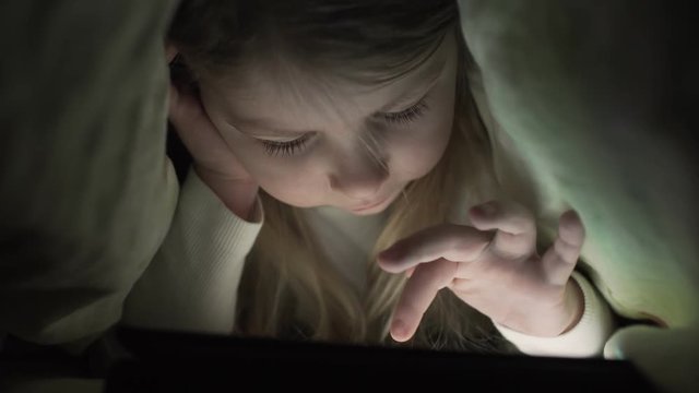 Young girl hiding under duvet to use digital tablet device late after bedtime. Little girl with tablet under blanket. Kid at night with tablet. Little girl with tablet in the bedroom.