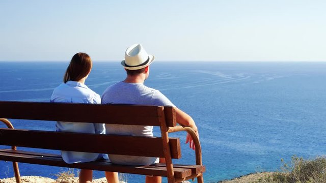 Young couple in love sitting relaxing on bench at edge of cliff. Husband in hat and wife on honeymoon enjoying breathtaking view of blue Mediterranean sea. Concept of freedom and love